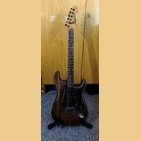 Elektrická kytara s tmavě hnědým dřevěným korpusem a hnedým hmatníkem. Kytara má tmavý vzorovaný pickguard a na hlavě je viditelné logo Fender spolu…