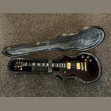 Tmavě hnědá elektrická kytara v otevřeném černém tvrdém pouzdře. Na hlavě nástroje je bílé logo Gibson. Kytara má dva humbuckery s krémovými kryty, z…