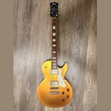Zlatá elektrická kytara typu Les Paul s dvěma humbuckery a bílým plechtrátem. Na hlavě je vidět logo Cort. Kytara má čtyři ovládací knoflíky a hриф s…