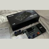 Na fotografii je kytarový procesor NUX MG-30 v černé barvě, doprovázený originální krabicí a dokumentací. Na přístroji je vidět nápis MG-30 VERSATILE…