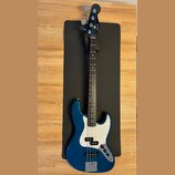 Modrá elektrická basová kytara s bílým perleťovým pickguardem. Na hlavě hmatníku je viditelné logo Fender. Nástroj má čtyři struny a kový most. Povrc…