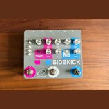Šedý efektový pedál s nápisem SIDEKICK a logem crazy tube circuits. Zařízení obsahuje několik bílých a fialových ovládacích knoflíků, tři přepínače v…