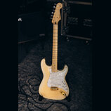 Elektrická kytara s bílým tělem a světlou hnedou hmatníkem. Na hlavě je viditelné logo Fender. Kytara má perleťový pickguard a zlaté kování ladících…