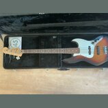Elektrická basová kytara s tělem v sunburst provedení a bílým pickguardem, umístěná v černém tvrdém kufru. Na hlavě nástroje je viditelné logo Fender…