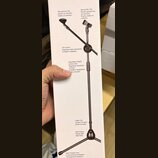 Fotografie zachycuje bílé balení s obrázkem černého mikrofonního stojanu. Na krabici jsou anglicky, polsky a česky popsané části stojanu: Microphone…