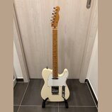 Krémová elektrická kytara typu Telecaster s javorovým hnedým hmatníkem a bílým pickguardem. Na hlavě nástroje je viditelné logo Fender. Kytara stojí…