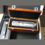 Stříbrný kondenzátorový mikrofon v dřevěném kufru s černým polstrováním. Na těle mikrofonu je viditelné kulaté logo se písmenem „S“. Kufřík má na pře…