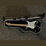 Černá elektrická kytara s bílým pickguardem v černém tvrdém kufru. Na hlavě kytary je viditelné logo Fender. Nástroj má světle hnědý javorový hmat a…