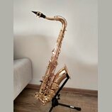 Zlatý saxofon s černým mouthpiecem, umístěný na černém stojanu. Nástroj má lesklý povrch bez viditelných poškzení. Na fotce nejsou žádné čitelné znač…