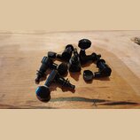 Sada černých ladicích kolíků z kovu. Kolíky mají hladký povrch a jsou rozházeny na dřevěném podkladu. Na dílech nejsou žádné viditelné značky ani náp…