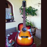 Akustická kytara s cutawayem v červeno-žlutém sunburst provedení. Na hlavě je viditelné modré logo s ptačím motivem. Hриф má tmavý dřevěný povrch a b…