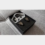 V černé krabičce s pěnovou vložkou jsou umístěny dva černý sluchátka in-ear a k nim příslušný stříbrný opletený kabel. Na sluchátkách jsou viditelné…