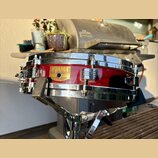 Červený snare drum s chromovanými obručmi a ladicími šrouby. Na korpusu je zlatý nápis TAMA a štítek s textem SNARE DRUM, LEGAL No. 200, MADE IN JAPA…