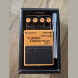Kytarový efekt v oranžovém provedení s černým pedálem, umístěný v černé krabici značky BOSS. Na přístroji jsou nápisy TURBO Distortion DS-2, OUTPUT,…