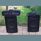 Dva černé reproduktory značky ALESIS s kovovými mřížkami. Na levém reproduktoru je umístěný černý mixážní pult s nápisy ALESIS a PAM. Na pultu jsou v…