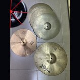 Fotografie zobrazuje sadu činelů v různých barvách a stavech. Jeden činel je černý s nápisem Zildjian, další jsou v přirozeném mosazném a stříbřitých…