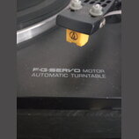 Detailní záběr na černý gramofon s nápisem F·G·SERVO MOTOR AUTOMATIC TURNTABLE. Na raménku je nasazen žlutý hloubkový systém s logem Audio-Technica.…