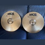 Dvě mosazné činely s koncentrickým rýhováním, položené na modrém koženém povrchu. Oba mají černý nápis MCS a logo MEINL. Na levém činele je drobný ná…