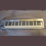 Stříbrný MIDI kontrolér s klaviaturní klávesou. Na horním panelu je zlatý pruh a nápis M-AUDIO KEYSTATION 61. V levé části jsou umístěny ovládací kno…