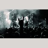 Černobílá fotografie zachycující dav lidí na koncertě. V popředí je vidět silueta natažené ruky s gestem „rocková roha“. V pozadí je osvětlené pódium…