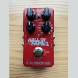 Červený kytarový efekt s nápisem HALL OF FAME 2 a logem tc electronic. Na horní části jsou čtyři černé ovládací knoflíky a nápisy PRE-DELAY (SHORT, L…