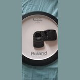 Bílý elektronický činel s černým gumovým okrajem a černým plastovým úchytem v centru. Na povrchu jsou nápisy V-Drums, CY-13R Ride a Roland. Dole je u…