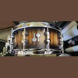 Fotografie zobrazuje snare drum s dřevěným korpusem v hnědém přirozeném odstínu s viditnou masivním kresbou dřeva. Uprostřed korpusu je umístěno stří…