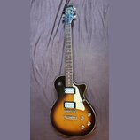 Elektrická kytara s trupem v tmavě hnědé a žluté barvě (sunburst) a bílým pickguardem. Na hlavě hmatníku je nápis Samick a pod ním logo ve tvaru štít…