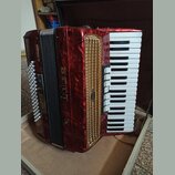 Červená akordeon s klaviatura a zlatou mřížkou. Na boku je svisle napsaný bílý nápis DELI a pod ním písmeno V. Na zlaté mřížce je malý oválný štítek…