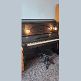Černé pianino s doprovodnou kulatou židličkou. Nad klávesnicí je v centru umístěn nápis ROCK & RIGELSY a nad ním oválný dekorativní prvek. Nástroj je…
