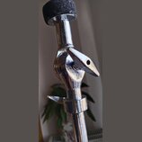 Detail chromovaného spojovacího kloubu stativu s černým knoflíkem nahoře. Na kovové části je vyryté logo připomínající stylizované písmeno S. Povrch…