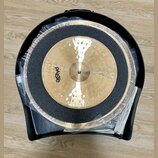 Dvě mosazné činely v černém plastovém kufru, přikryté průhlednou fólií. Na horním činele je viditelné logo Paiste. Mezi činely je umístěna černá tlum…
