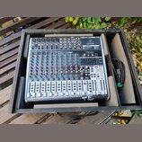 Stříbrný mixážní pult značky behringer s nápisem XENYX QX1832USB umístěný v černé dřevěné bedně. Zařízení obsahuje numerous červené a šedé otočné kno…