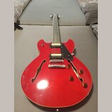 Červená polodublová elektrická kytara s dvěma humbuckery a bílými rámečky. Nástroj má hnědé ovládací knoflíky a černý plechtr zaštitu. Na kytáře nejs…