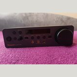 Tmavě šedý audio interface s digitálním displejem a velkým ovládacím knoflíkem vpravo. Na předním panelu je nápis Sound BLASTER a několik tlačítek s…