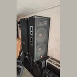Dvě velké černé reproduktorové bedny s kovovými mřížkami a plastovými rohy. Na pravé bedně je v dolní části viditelné oranžové logo JBL. Pod nimi jso…