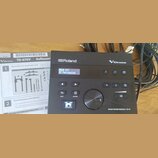 Černý elektronický modul s nápisy Roland a V-Drums. Na displeji je text DRUM KIT 25 Percussion a v pravém dolním rohu nápis DRUM SOUND MODULE TD-07.…