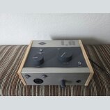Šedý audio procesor v dřevěném rámu. Na horním panelu je logo UA, nápisy INPUT a OUTPUT s LED indikátory, přepínač s označením VOC, GTR, FAST, OFF a…