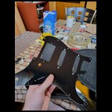 Černý plastový pickguard pro elektrickou kytaru s třemi výřezy pro snímače. Na každém z nich je umístěn černý kryt s bílým nápisem ETIG. Povrch pickg…