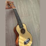 Dřevěný ukulele s přírodní barvou těla a hnědým krčkem. Na hlavě je vidět nápis CASUAL. Nástroj má čtyři struny, tmavý hmatník a černý lem kolem ozvu…