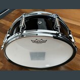 Černý buben s chromovaným obručováním a mechanismy. Na bílém koženu je vidět logo REMO a nápis SILENTSTROKE™ MADE IN U.S.A. Povrch nástroje je glossy…
