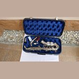Stříbrný saxofon v černém kufru s modrým polstrováním. K nástroji je přiložen h mouthpiece v plastovém obalu a červený popruh. Na těle nástroje nejso…
