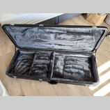 Otevřený černý kufr na hudební nástroj s vnitřním polstrováním z šedého plyšového materiálu. V dolní části jsou vidět tři oddělené přihrádky pro ulož…