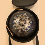 Kovový perkusní nástroj typu steel tongue drum v tmavě modré barvě s geometrickým bílým vzorem ve středu. Nástroj je umístěn v černé přepravní brašně…