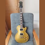 Zlatá elektrická kytara typu Les Paul s dvěma humbuckery a čtyřmi ovládacími knoflíky. Na hlavě je bílý nápis Epiphone a zlatý nápis Les Paul. Kytara…