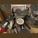 Elektronická bicí souprava značky Roland s černými pady a činely. Na module je nápis Roland TD-5 V-Drums. Centrální pad má bílý povrch s logem Roland…