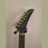 Fotografie zachycuje hlavu elektrické kytary v černé barvě. Na špičce hlavy je bílý nápis Epiphone. Nástroj má šest zlatých ladiček a hnedík z tmavéh…