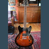 Polodupatá elektrická kytara s tělem v barvě sunburst a černým pickguardem. Na hlavě je viditelné logo Epiphone. Nástroj má dva humbuckery, tři kontr…