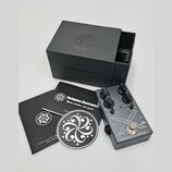 Kytarový efekt pedál stříbrné barvy s černými ovládacími knoflíky a nápisy MICROTUBES X, Darkglass Electronics - MADE IN FINLAND. Vedle pedálu je čer…