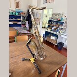 Stříbrný saxofon umístěný na černém stojanu značky HERCULES. Nástroj vykazuje viditelné opotřebení povrchu, zaprášení a místní oxidaci kovu. Na těle…